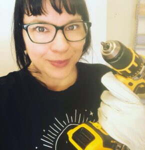 Ducky grinning at the camera while wearing work gloves and holing a drill.